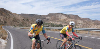 Two cyclists, a man and a woman, bicycling on an empty stretch of highway on the Baja California Peninsula wearing yellow and black cycling clothing and bicycle helmets. The man gives the photographer a thumbs-up with his left hand